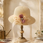 A cream-colored woven sun hat with a wide brim, decorated with a large cream and dusty pink fabric flower with a jeweled center, displayed on a vintage-style wooden stand. The scene is styled with a brass watering can, a brown leather journal, dried lavender, a white rose in a teacup, and a lace runner against a rustic stone wall background.