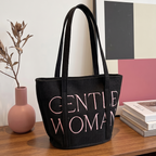 Black tote bag with 'GENTLE WOMAN' text on a wooden surface with books and a vase.