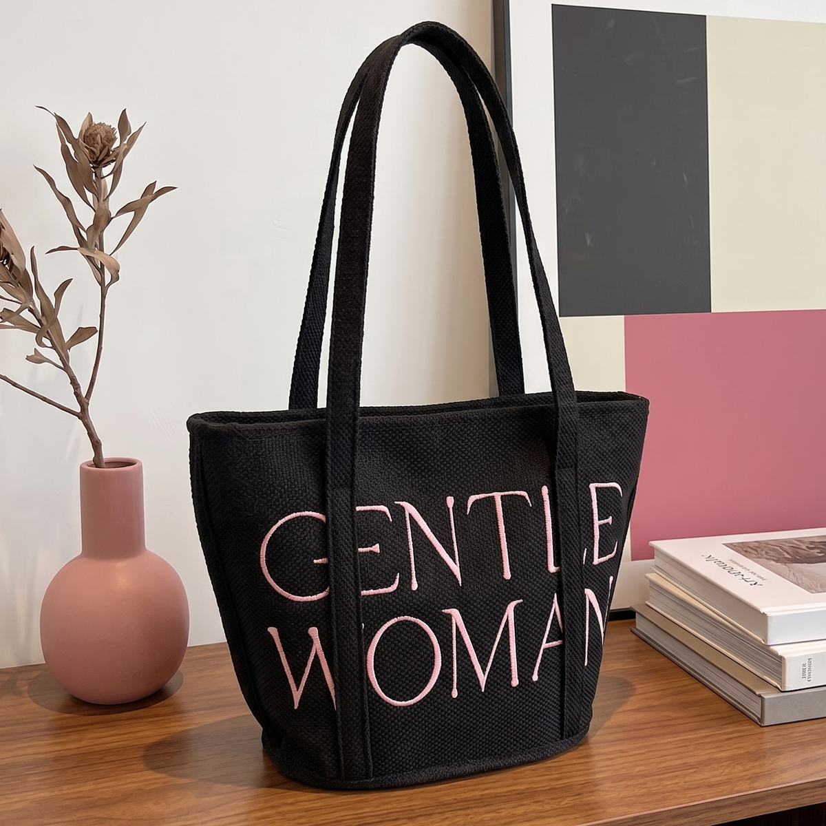 Black tote bag with 'GENTLE WOMAN' text on a wooden surface with books and a vase.